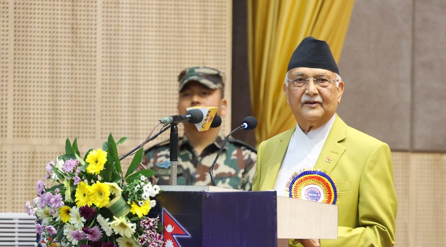 KP Sharma Oli delivers a speech on National ICT Day in Kathmandu, with a floral podium and a soldier in the background.
