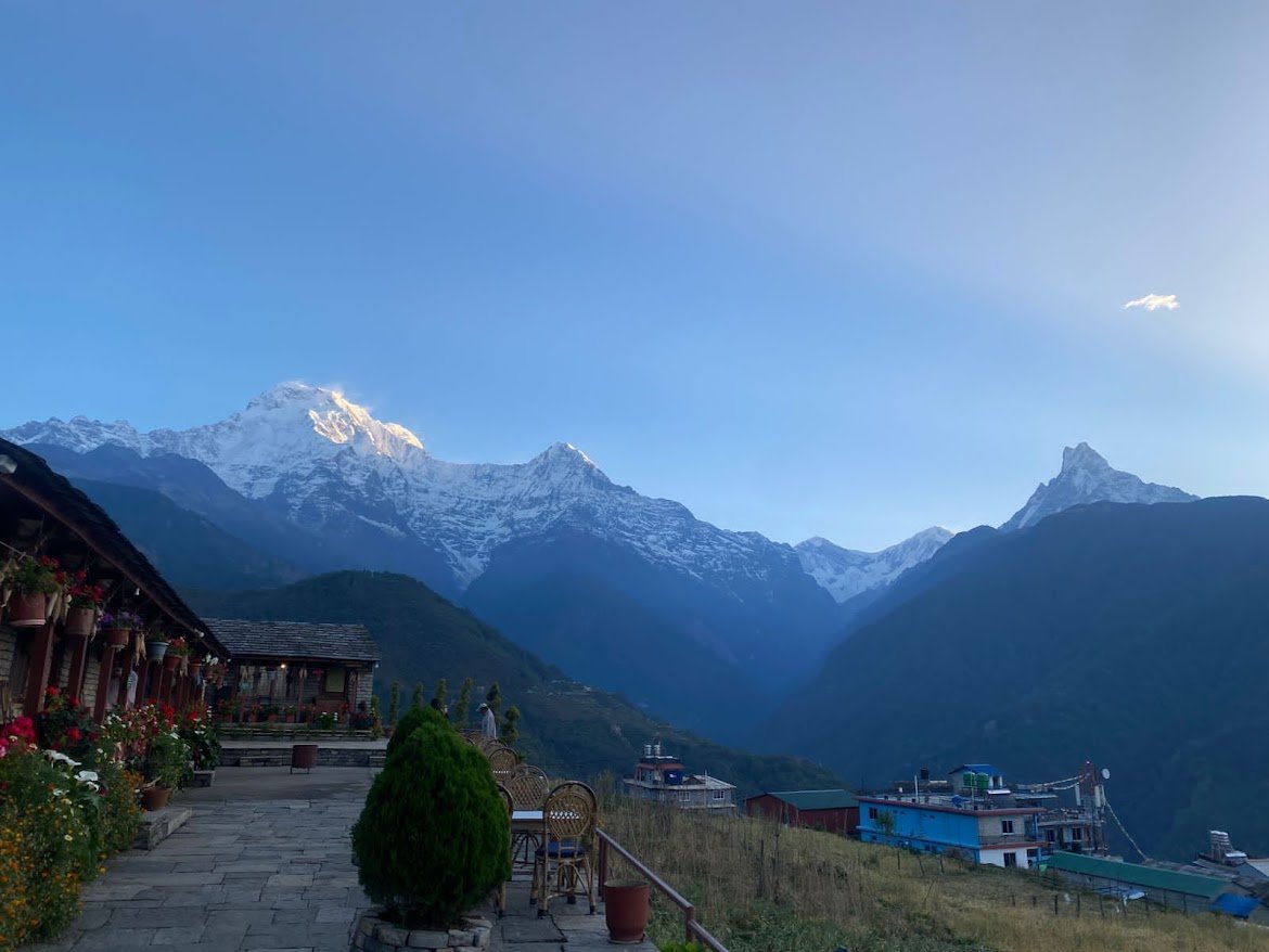 Scenic view of Annapurna mountain range at sunrise from Gurung Cottage Nepal, with flowers, cottages, and Himalayan peaks in the background.