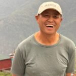 Kisam Gurung standing outdoors in Ghandruk, smiling in front of a mountain backdrop, wearing a grey shirt and a cap.
