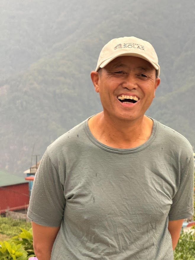Kisam Gurung standing outdoors in Ghandruk, smiling in front of a mountain backdrop, wearing a grey shirt and a cap.