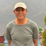 Kisam Gurung standing outdoors in Ghandruk, smiling in front of a mountain backdrop, wearing a grey shirt and a cap.