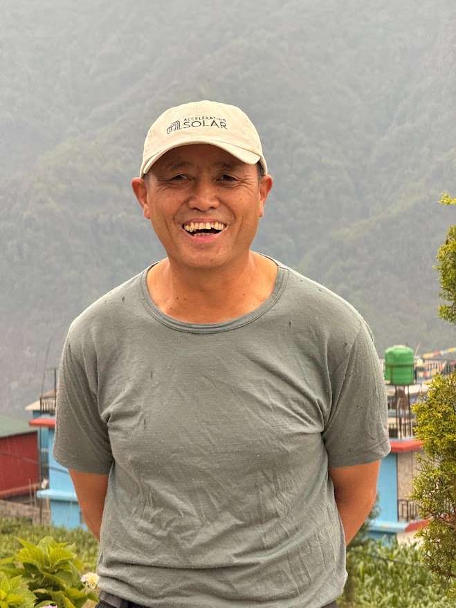 Kisam Gurung standing outdoors in Ghandruk, smiling in front of a mountain backdrop, wearing a grey shirt and a cap.