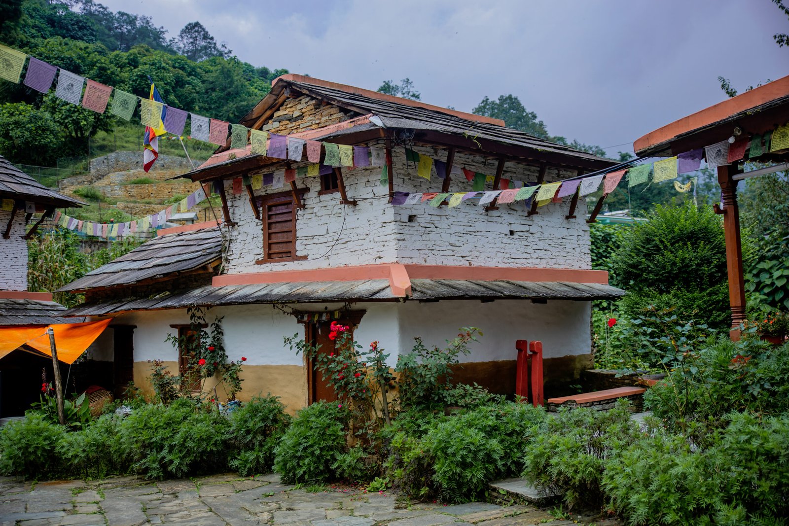 Rustic Gurung house in Ghandruk village, Nepal, with stone roofing, prayer flags, and flowering plants, photographed by Subash Thapa Magar.