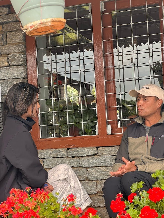 Kisam Gurung, wearing a beige cap and outdoor jacket, sits and talks with Sushmita Baniya, who is dressed in a black jacket and patterned trousers. They are seated on a stone bench in front of a window with metal bars, surrounded by red flowers and potted plants.2/2 