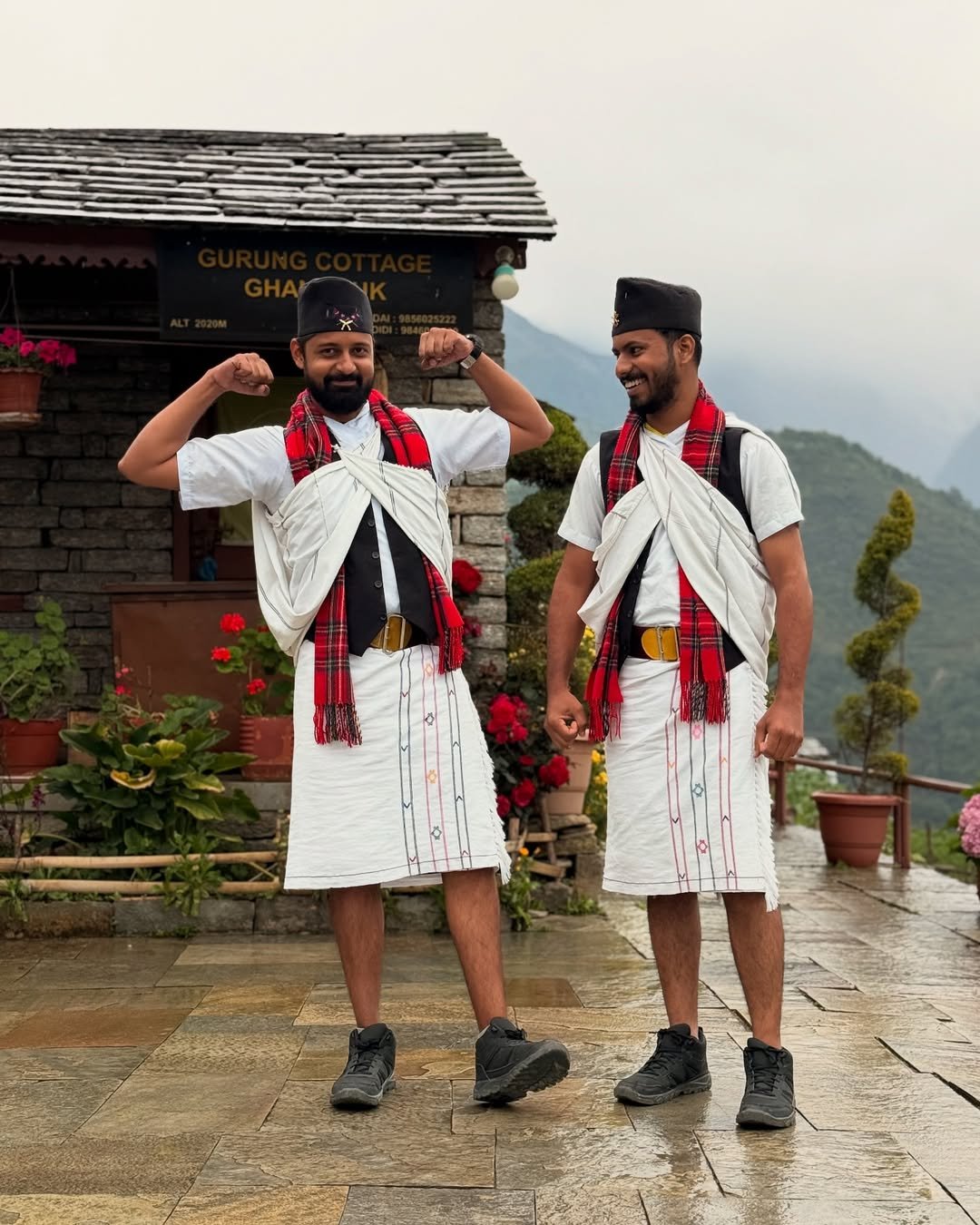 Two men dressed in traditional white Gurung outfits with red checked scarves and black topis stand smiling in front of Gurung Cottage in Ghandruk. The man on the left flexes his arms while the man on the right laughs. The background features stone buildings, vibrant flowers, and misty mountains.